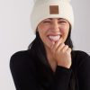 A person with long dark hair is smiling broadly with eyes closed, wearing a white Baylor Beanie - Sweet Cream by Woolx featuring a brown leather patch on the front and a black shirt. Their right hand is brought up to their mouth, and they have a joyful expression.