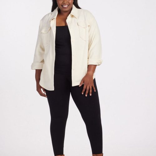 A person stands smiling in a Sawyer Shacket by Woolx, in Sweet Cream color, layered over a cream-colored button-up shirt, paired with black leggings and brown ankle boots, posed against a plain white background.