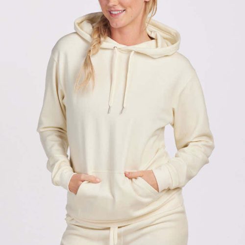 Dressed in a cream Woolx Avery Hoodie from the Stocking Stuffer Final Sale Deals, paired with matching sweatpants, an individual stands against a plain background. With their hair styled in a side braid, they smile while placing their hands in the front pocket of the hoodie.