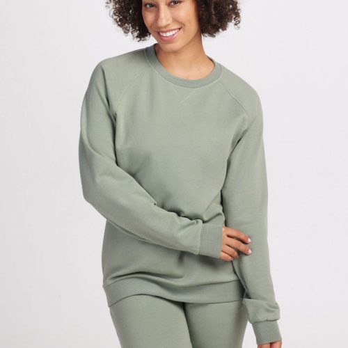 A person with curly hair smiles while wearing a sustainable Wool Aire™ Bailey sweatshirt and matching pants from Woolx. They stand against a plain white background, one hand on their hip and the other resting on their leg.