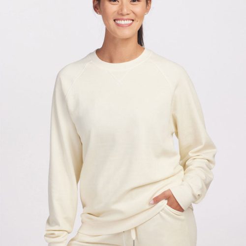 A person with long hair smiles while wearing a cream-colored Woolx Wool Aire™ Bailey sweatshirt and matching pants made from sustainable Merino wool against a plain white background. One hand is in their pocket, and the other hangs by their side.