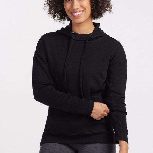 A person with curly hair is smiling while wearing a cozy Callie Hoodie - Black from Woolx and gray leggings. They are standing against a plain white background with one hand touching their hip and the other resting on their stomach. | Tori is 5'7", wearing a size S