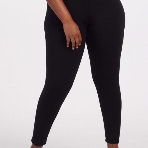 A person wearing Woolx's Stella Tall leggings in black stands against a plain white background. They are visible from the waist down, with their right hand resting on their thigh, highlighting the sleek design of these cozy baselayer leggings paired with black sneakers.