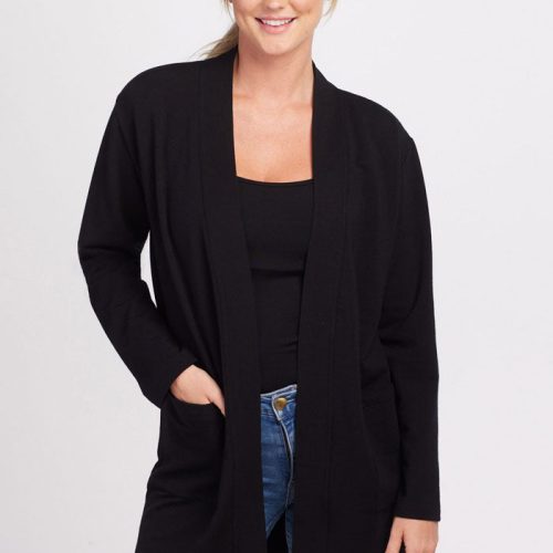 A person with blond hair is smiling at the camera, standing against a plain white background. They are wearing the Woolx Kinsley Cardigan in black over a matching black top and blue jeans. One hand is placed in the pocket of the cardigan, while the other hand rests by their side. | Karly is 5'10", wearing a size S