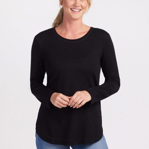 A woman with blonde hair tied back in a ponytail is smiling at the camera. She is wearing the Woolx Blair Tunic - Black, an ultra-cozy, long-sleeve top made from Australian Merino Wool, paired with light blue jeans. Her hands are clasped together in front of her against a plain white background. | Karly is 5'10', wearing a size S