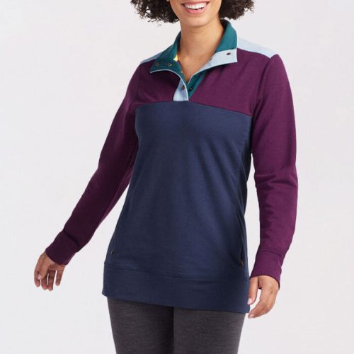 A person with curly hair smiles while wearing the Charlie Pullover from Woolx in a winter navy color, featuring a navy and purple color block design with a teal collar. Made from DuraLite Merino wool, they pair it with gray pants while standing against a plain white background.