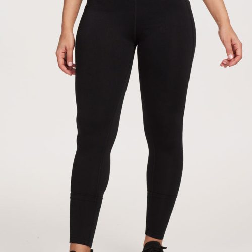 A person stands against a plain background, wearing Woolx's McKenna Pocket Leggings in black, paired with black sneakers featuring white soles. The leggings have side pockets and a high waistband for extra comfort, while the upper body is not visible.