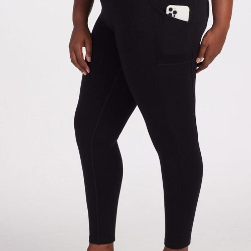 A person wearing the Piper Pocket Tall black leggings from Woolx, designed specifically for tall women, stands against a plain background. These high-waisted leggings have a snug fit with visible stitching details along the sides and feature a convenient side pocket with a smartphone in it, paired seamlessly with black sneakers.