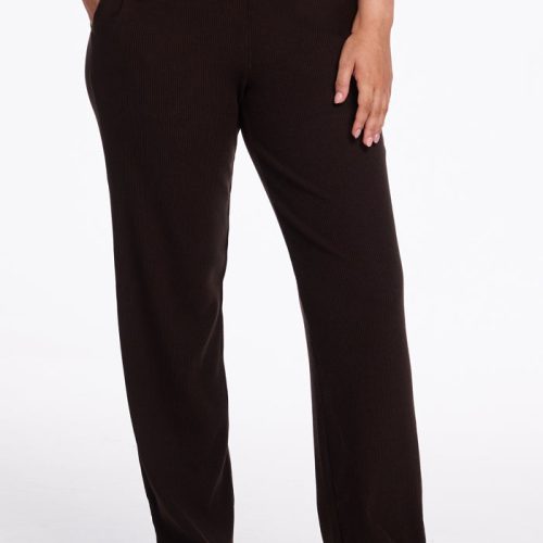 A person is standing with one hand in their pocket, modeling dark brown Luca Ribbed Pants - French Roast by Woolx, which feature a high-waisted, straight-leg fit. They are also wearing white sneakers. The background is plain white. | Sarah is 5'8", wearing a size S