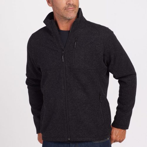 A man with short hair is wearing a Woolx Fairbanks Jacket in Carbon Black, featuring a zip-up design with a minimalistic aesthetic. Paired with jeans, he stands against a plain background, looking to the side with one hand in his pocket, highlighting the refined simplicity of his attire. | Lee is 6’, wearing a size M