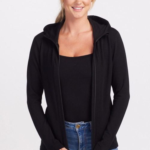 A person with long blonde hair is smiling while wearing the Woolx Black Zoey Zip Up Hoodie over a black top made of Australian Merino Wool and light blue jeans. The individual is posing with hands resting on the hips against a plain white background, showcasing perfect layering. | Karly is 5'10", wearing a size S