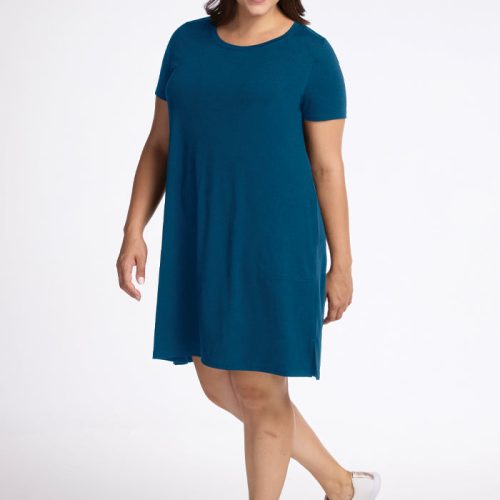 A person with long dark hair is smiling and wearing the Woolx Georgie Dress in Real Teal and white sneakers. They stand with one leg slightly raised and hand on hip against a plain white background.